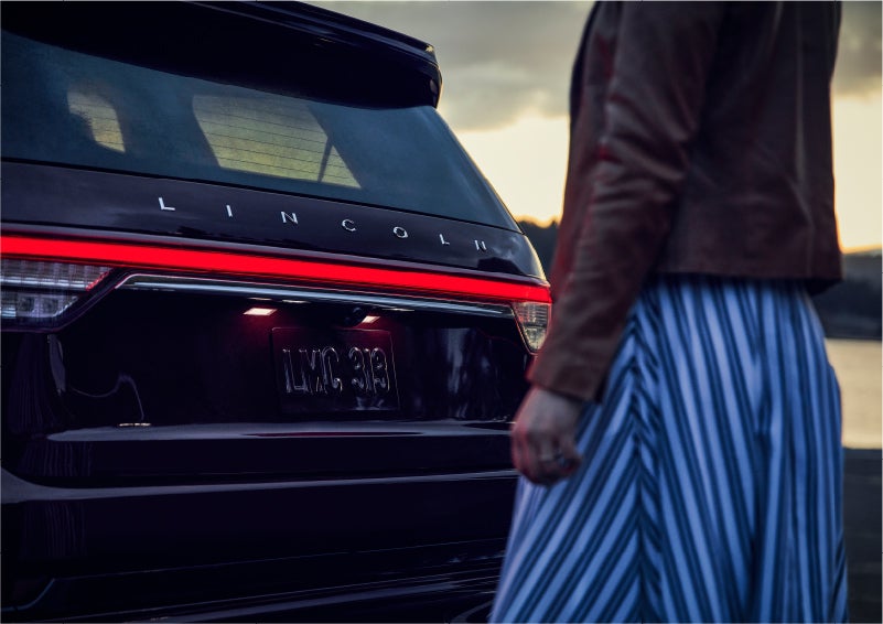 A person is shown near the rear of a 2023 Lincoln Aviator® SUV as the Lincoln Embrace illuminates the rear lights | Sedano Lincoln in La Mesa CA