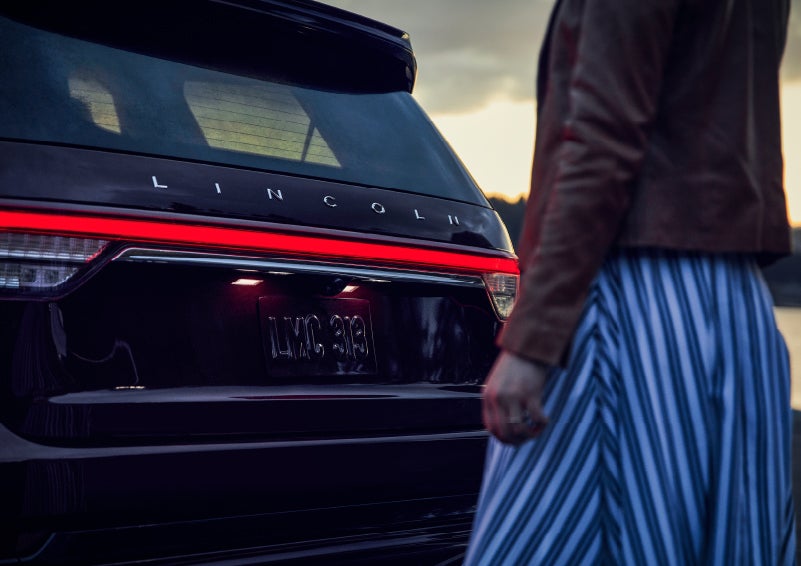 A person is shown near the rear of a 2024 Lincoln Aviator® SUV as the Lincoln Embrace illuminates the rear lights | Sedano Lincoln in La Mesa CA