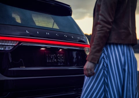 A person is shown near the rear of a 2024 Lincoln Aviator® SUV as the Lincoln Embrace illuminates the rear lights | Sedano Lincoln in La Mesa CA