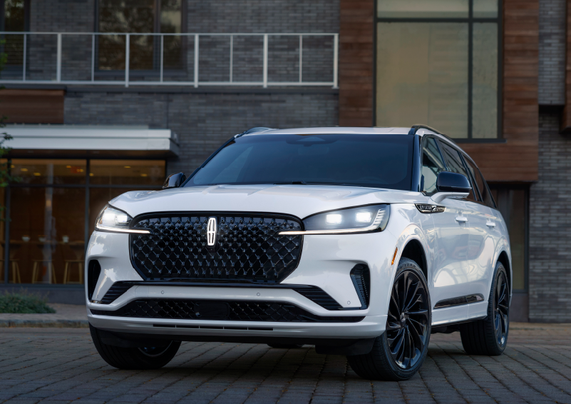 A white 2025 Lincoln Aviator® SUV is shown parked outside a modern building with gray brick and wood accents. | Sedano Lincoln in La Mesa CA